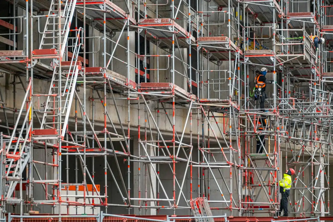 Fachada de edifício em obra inteiramente coberta por andaimes metálicos, com operários de capacete, colete refletivo e cinto de segurança sobre as plataformas