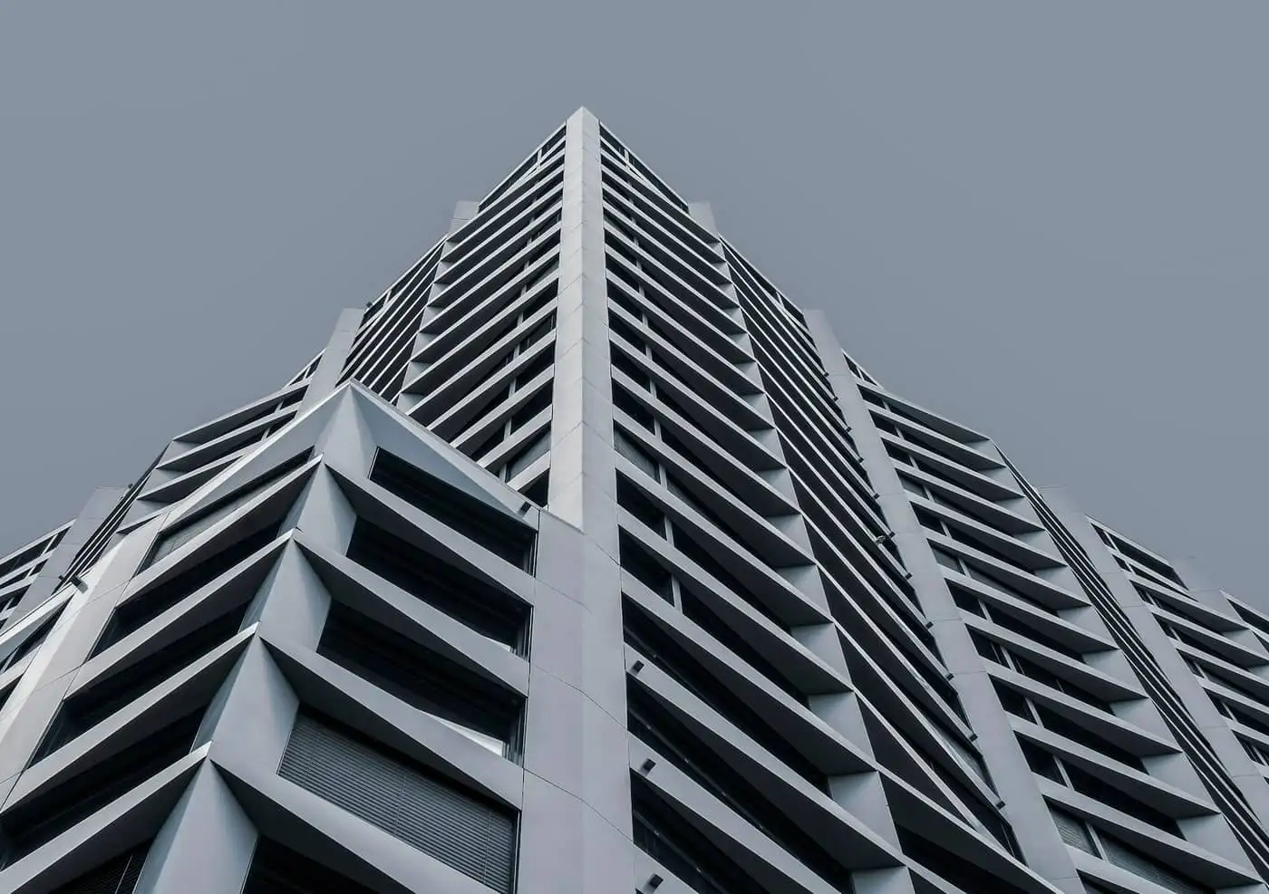 Edifício alto fotografado de baixo, com fachada de concreto claro em faixas horizontais e varandas recuadas, contra céu cinza uniforme