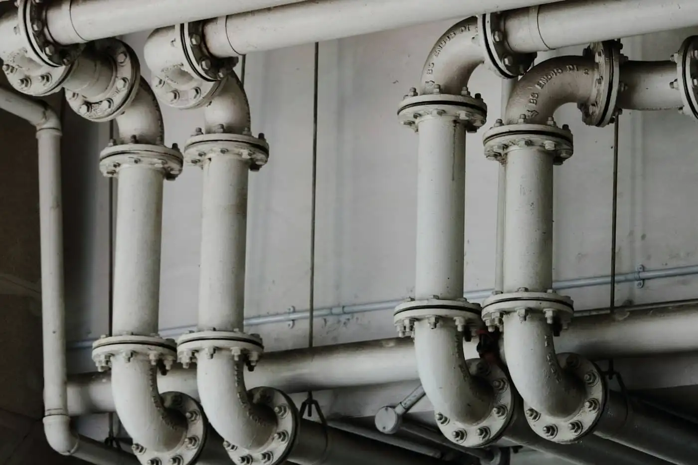 Detalhe de instalação hidráulica em tons de cinza, com tubos, curvas e conexões flangeadas fixados ao longo de uma parede