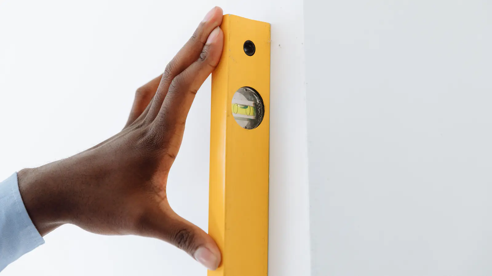 Mão apoia um nível de bolha amarelo numa parede branca, perto da quina, para conferir o prumo
