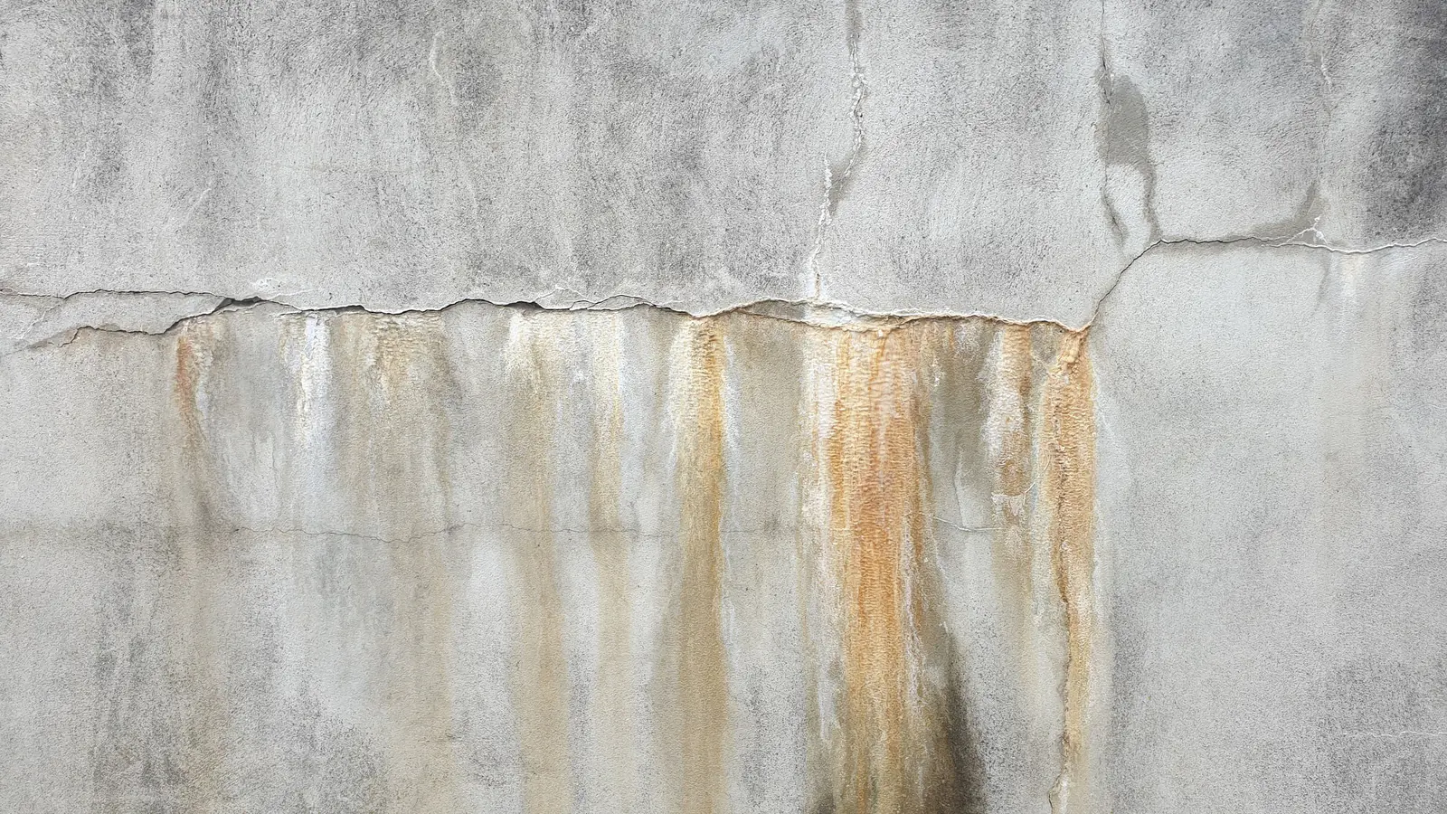 Parede de concreto com uma fissura horizontal e manchas de água escorrida logo abaixo, em tons de ferrugem e cinza